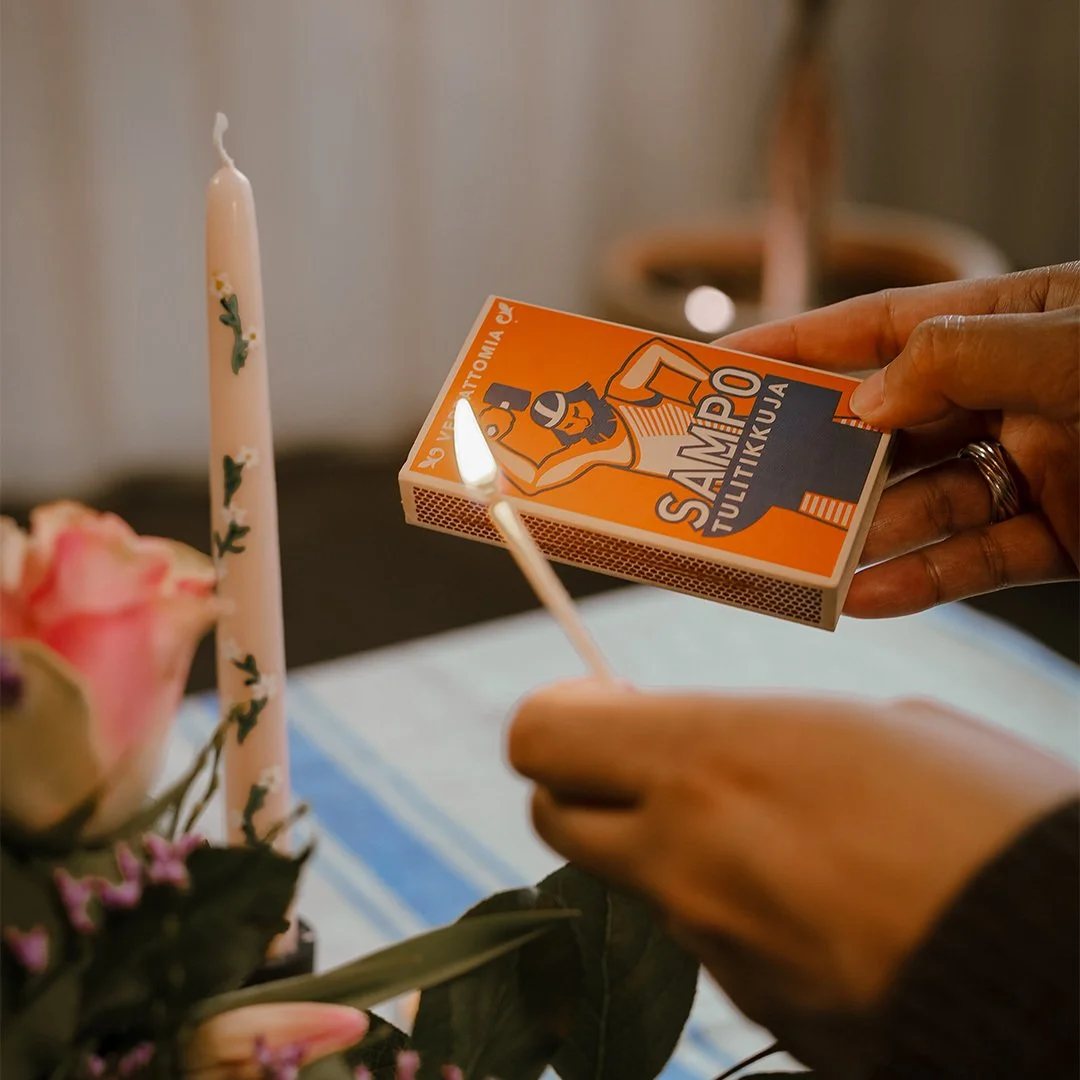 Person lighting a candle with a matchstick near a birthday cake and flowers.