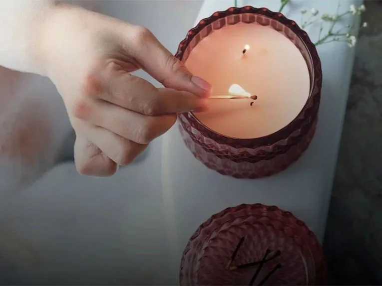 A person lighting a candle from a matchstick, with a second unlit candle on a white surface