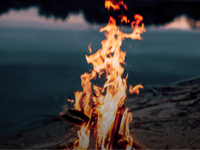 A campfire burning on a sandy beach near water, with dark clouds in the sky.