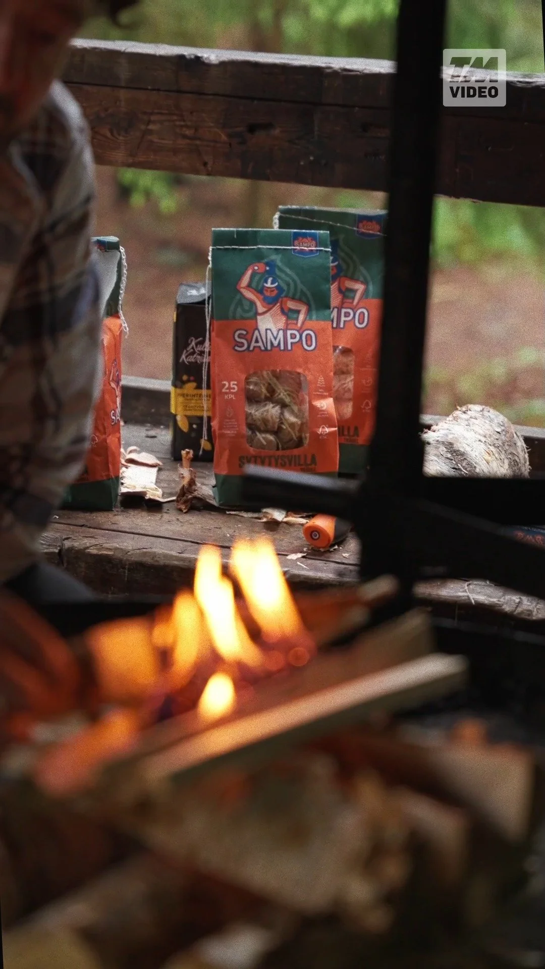 A wooden outdoor table stacked with packs of Sampo snack food, with a blurred figure on the left and a small fire in the foreground.