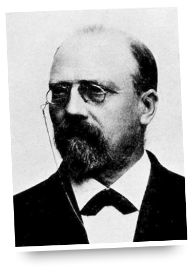 Svartvitt porträtt av en man med glasögon, skägg och mustasch, klädd i kostym och fluga.