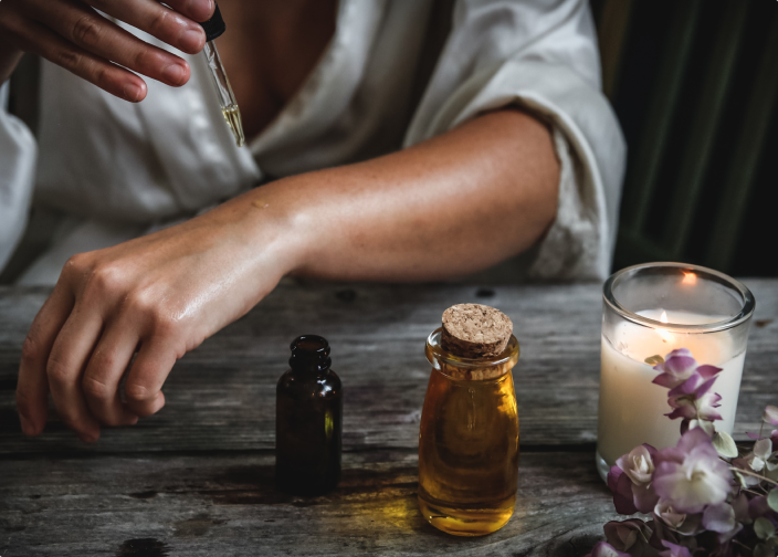 En persons hand som håller en pipett över en liten mörk flaska, med ytterligare flaskor och ett tänt ljus med blommor på ett träbord.