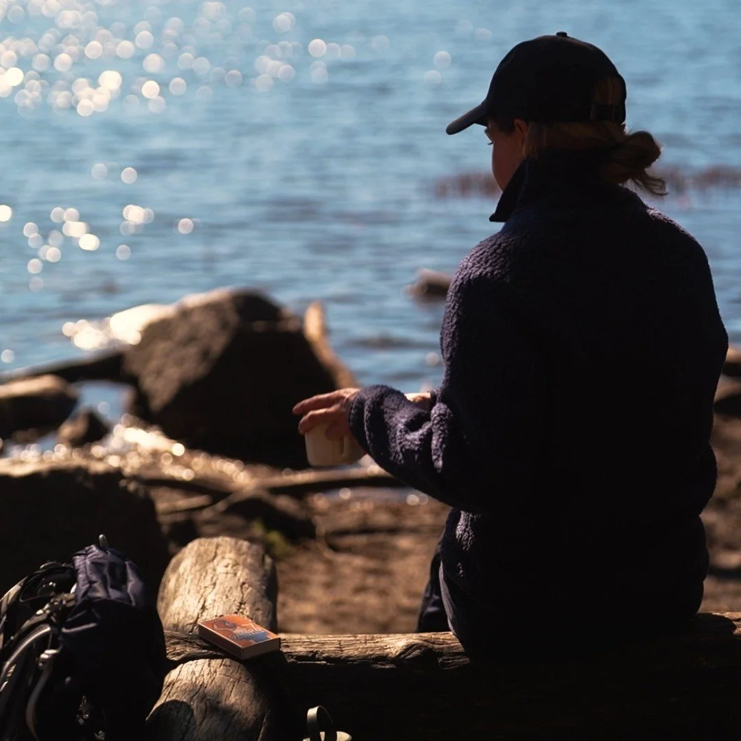Person with a cap sitting on a log near the water, holding a cup, with rocks and logs around and a bag and book nearby.