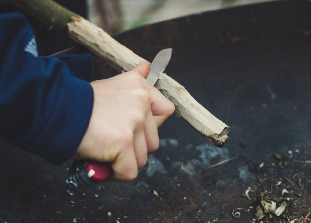 A person using a knife to carve or scrape a piece of wood over a black surface.