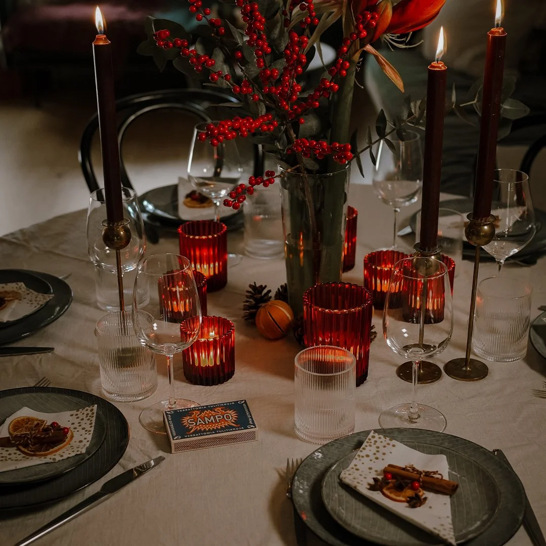A festive dinner table decorated with red candles, a large floral arrangement with red berries, and plates with napkins and food. The table has multiple glasses and is softly lit, creating a warm holiday atmosphere.