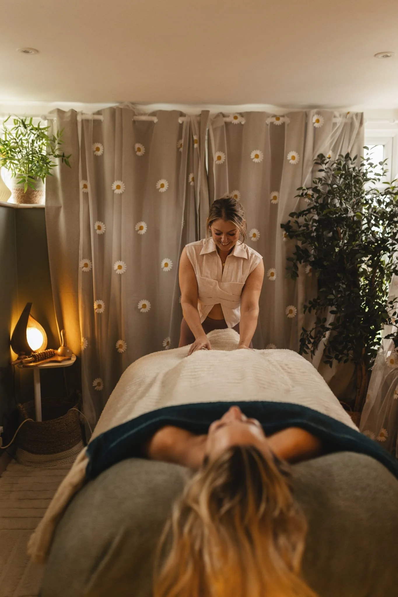 A woman receiving a massage in a cozy room with beige curtains decorated with daisies and potted plants, creating a relaxing ambiance.