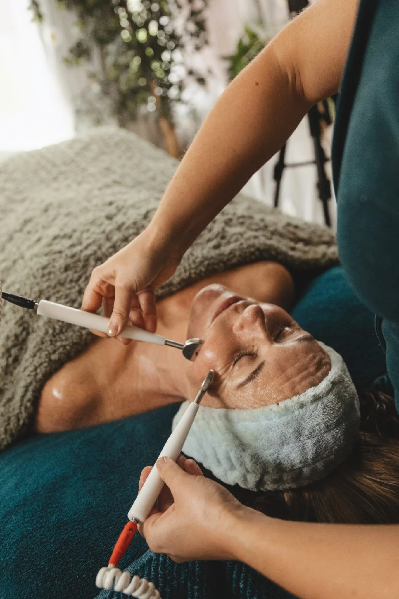 A person lying down on a bed with a towel wrapped around their head, receiving a facial treatment with multiple electrodes attached to their face, and a technician holding a device near the person's face.