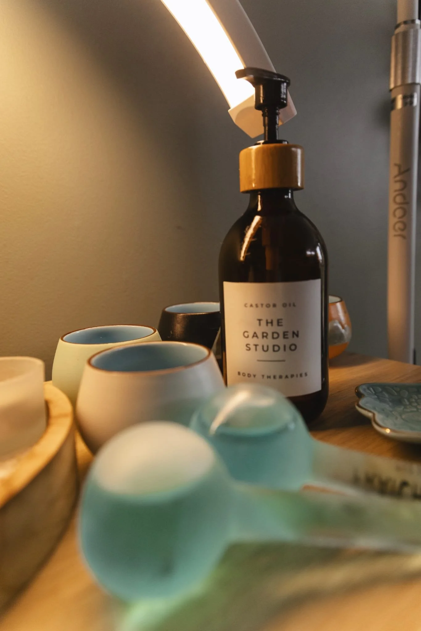 A desk scene with a dark amber bottle labeled 'Castor Oil, The Garden Studio, Body Therapies,' surrounded by small ceramic cups and bowls, with a lamp providing warm lighting.