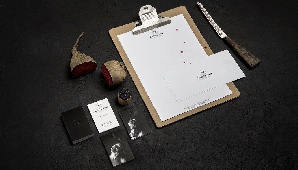 Corporate Design Geschäftsdrucksorten, auf einem schwarzen Hintergrund, daneben Karten, eine Metallklammer auf einer braunen Klemme, Rote Beete, ein Messer mit Holzgriff und Klingenmuster, sowie verschiedene Visitenkarten und eine Kreidetafel
