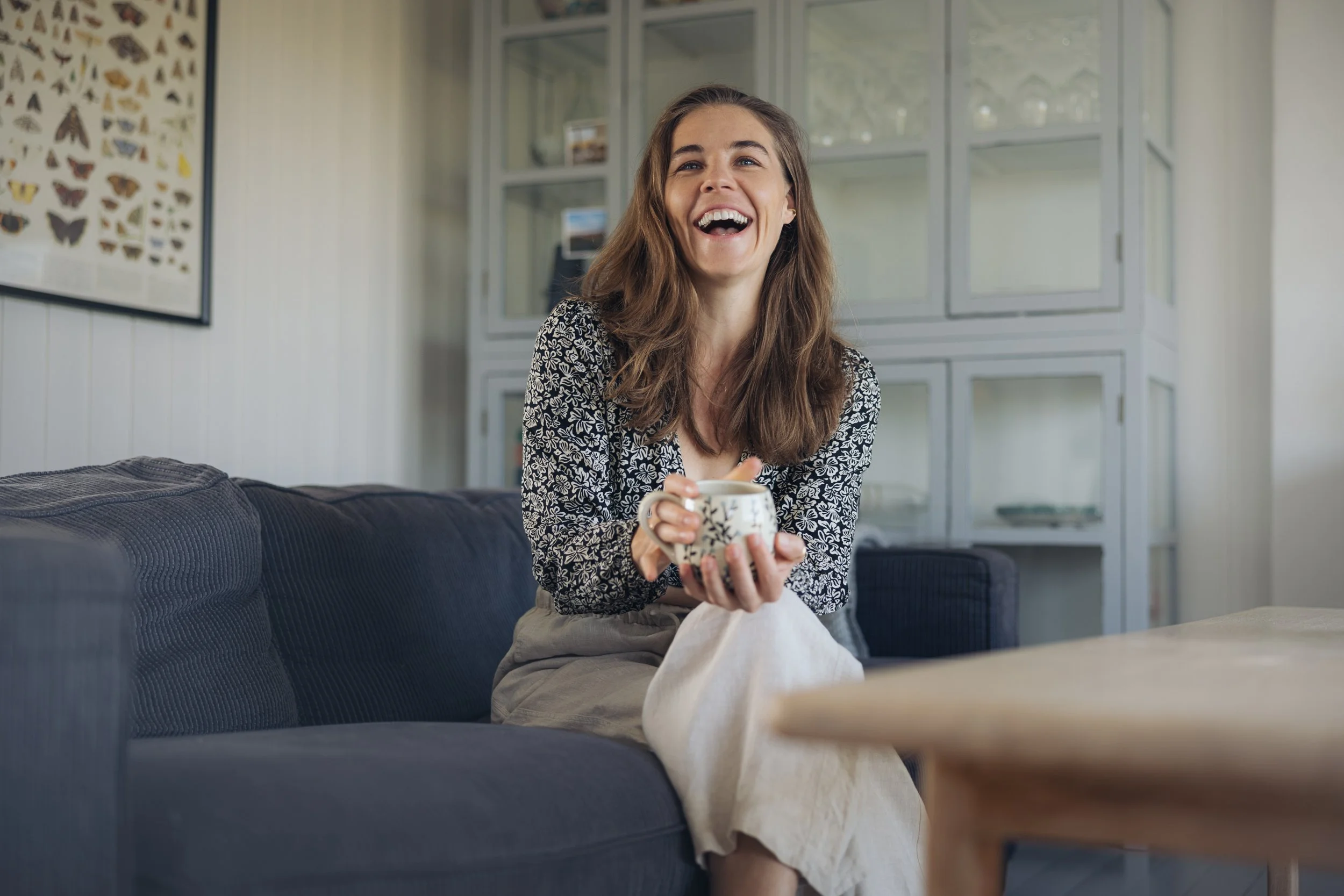 Eine lachende junge Frau mit braunen Haaren sitzt auf einem grauen Sofa und hält eine Kaffeetasse in den Händen, im Hintergrund ein Fenster und eine Wand mit einer ausgestopften Schmetterlingsausstellung.