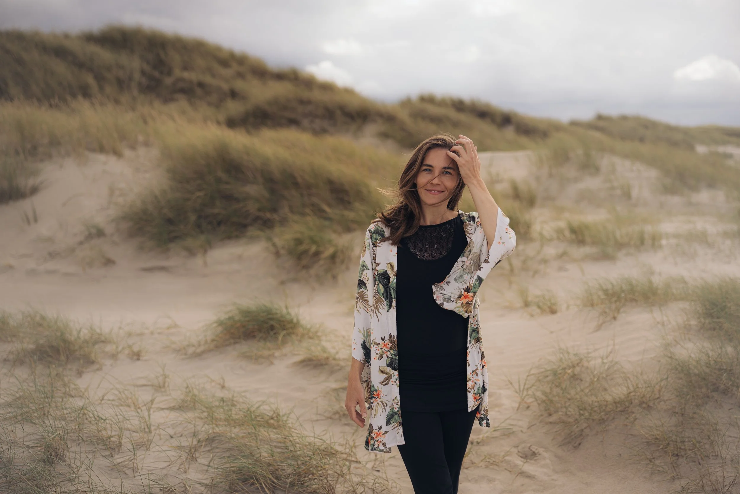 Eine Frau steht am Strand vor Dünen und Gräsern, trägt eine Blumenjacke und lächelt, während sie eine Hand in die Haare legt.