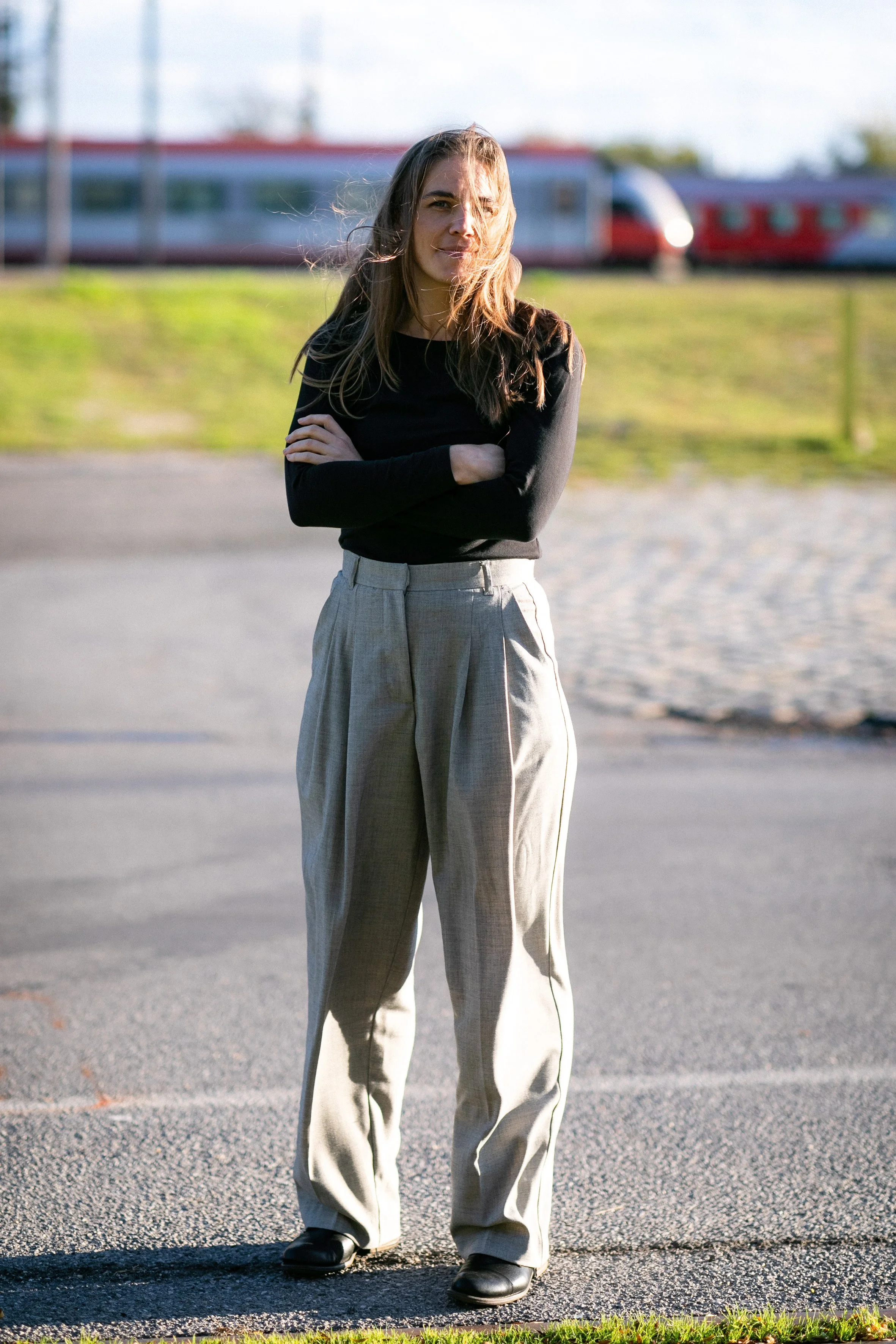 Junge Frau mit im Wind flatterndem Haar und schwarzen Schuhen steht mit verschränkten Armen auf einem Asphaltweg, im Hintergrund ein rot-weißes Fahrzeug und grüne Wiese in der Abendsonne.