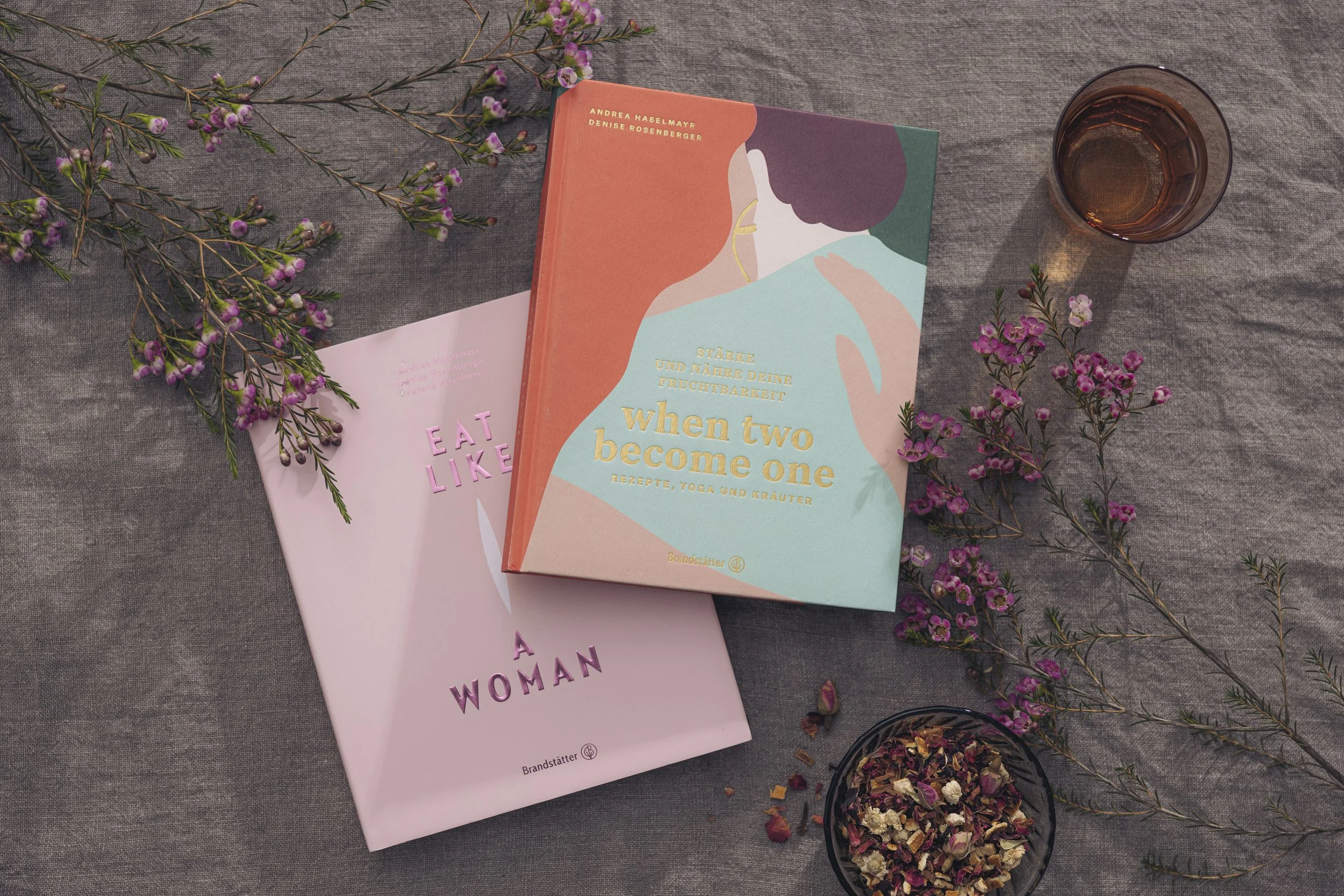Zwei Bücher, eines mit einem bunten Cover und einem weiblichen Porträt, liegen auf einem grauen Tuch. Es sind Zweige mit kleinen lila Blumen und eine Tasse mit Tee oder Kaffee sowie eine Schale mit getrockneten Blüten oder Kräutern daneben.