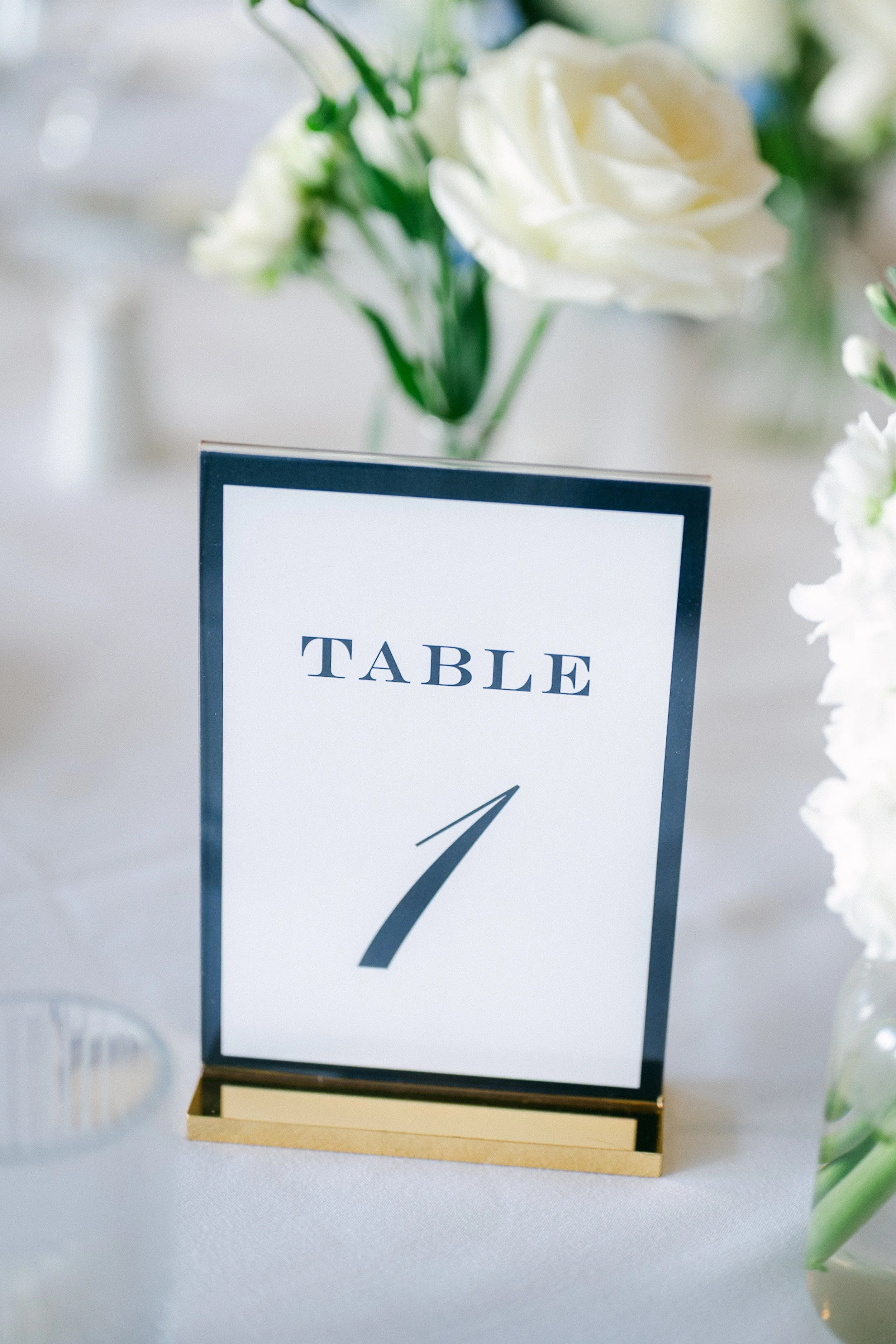 Table number sign marked 'Table 1' on a wedding or event reception table with white flowers in vases in the background.