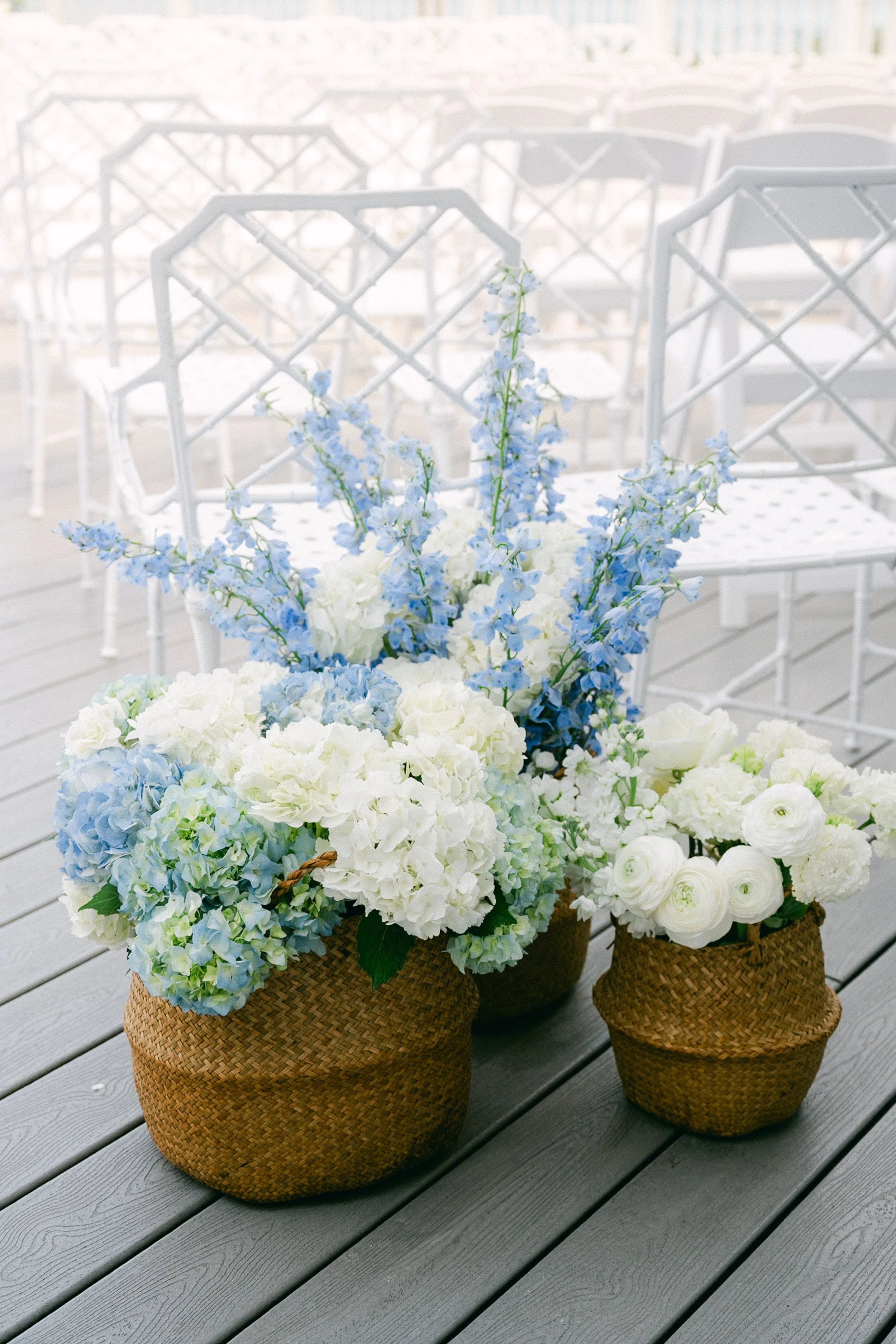 Basket of white and light blue flowers on a wooden table with white chairs in the background.