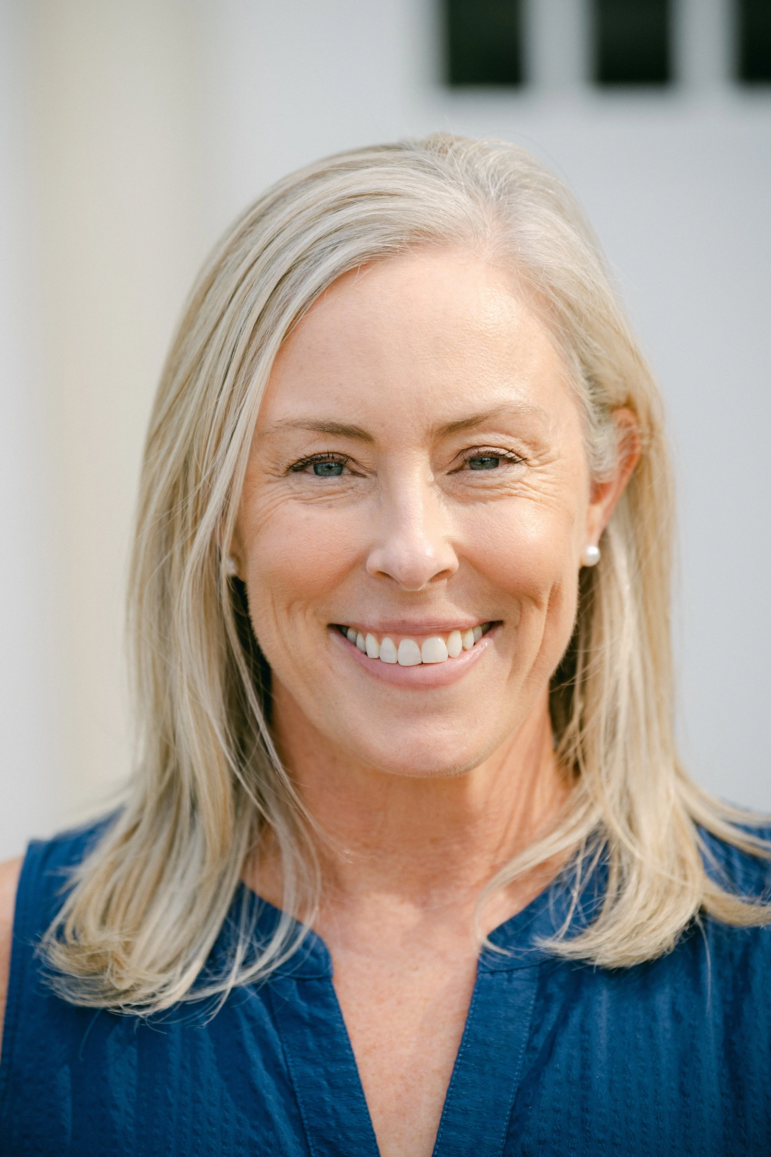 Smiling middle-aged woman with blonde hair, blue eyes, and pearl earrings, wearing a sleeveless dark blue top.