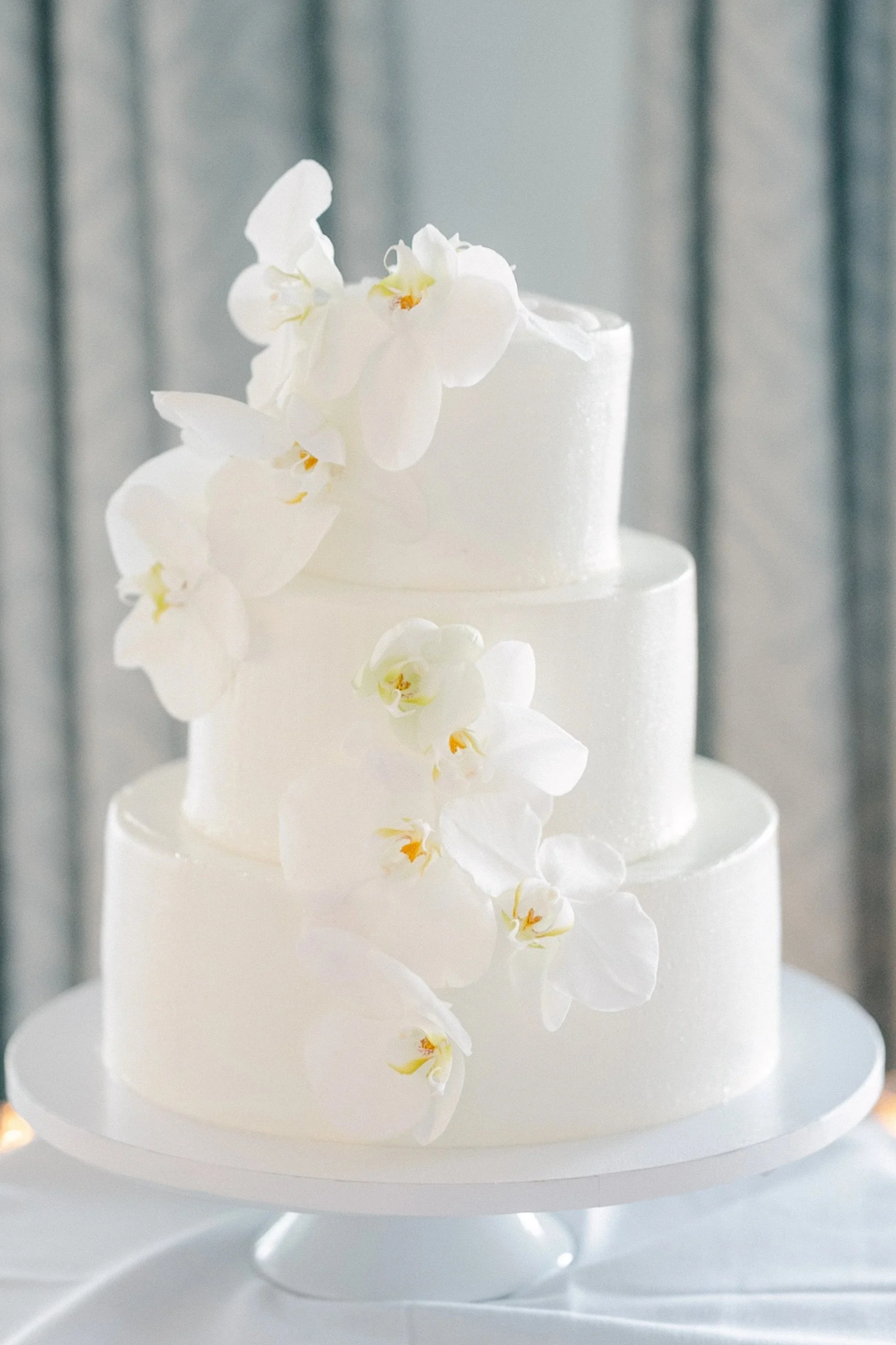 A three-tiered white wedding cake decorated with white orchids placed on a white cake stand.