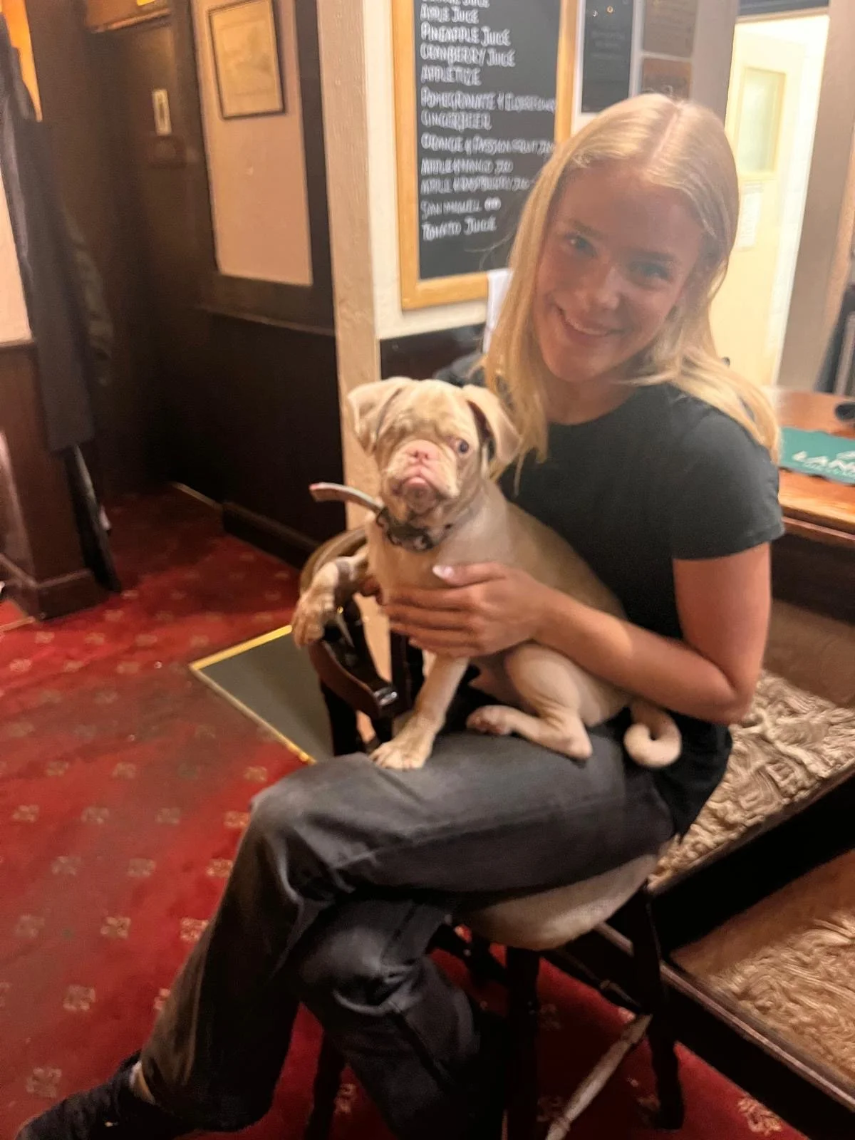 A smiling woman with blonde hair sitting on a chair while holding a puppy in her lap in a cozy restaurant or cafe with wooden decor and a menu board in the background.