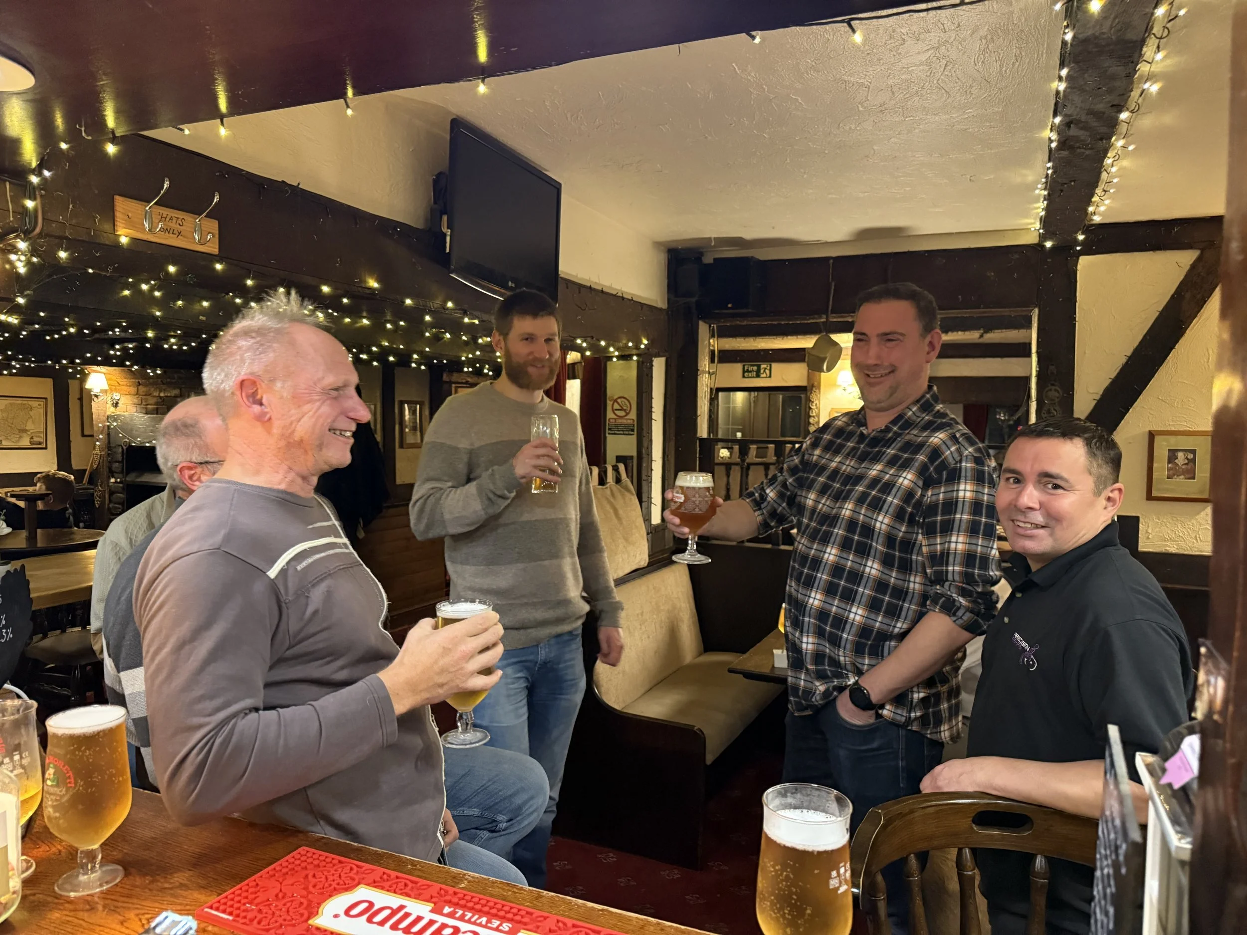 Group of five men inside a pub, holding glasses of beer, smiling and engaging in conversation. The setting features string lights and wooden beams.