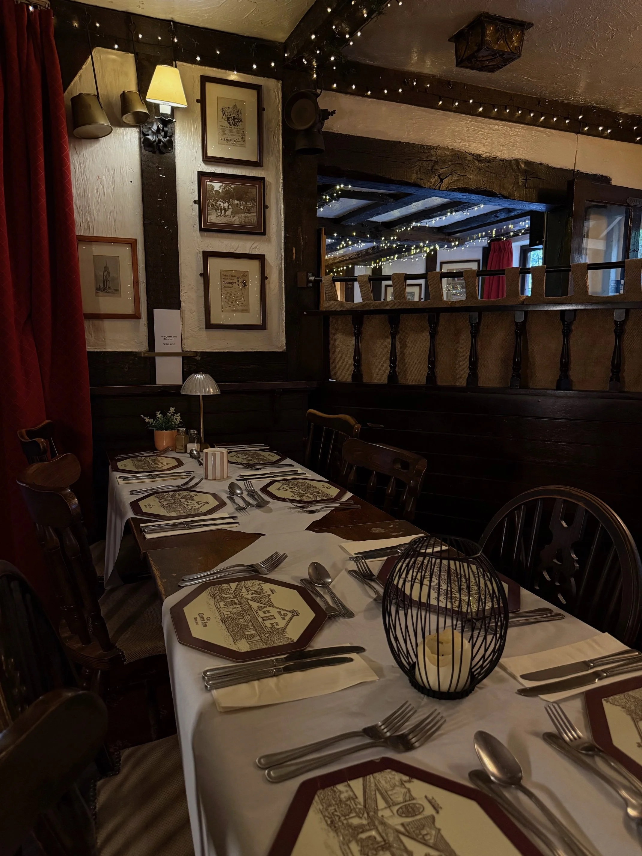 A cozy restaurant interior with a table set for a meal, featuring a white tablecloth, placemats with illustrations, silverware, and a small lamp. In the background, there are framed pictures on the wall, string lights on the ceiling, and a view into 