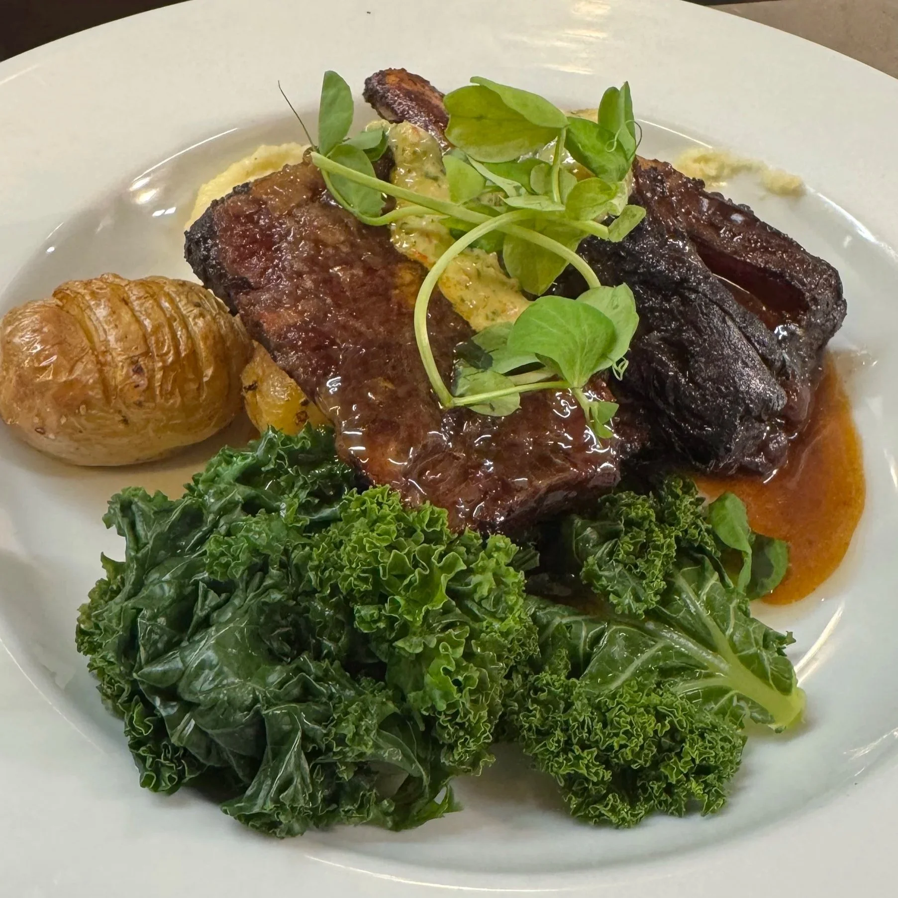 Plate of roasted beef ribs with gravy, garnished with green microgreens, served with a small roasted potato, kale, and spinach