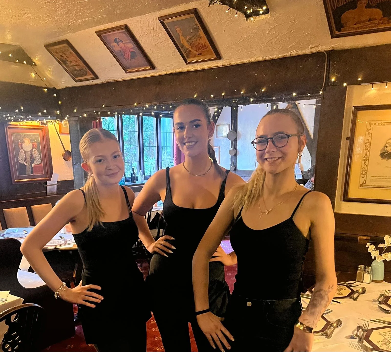 Three women in black dresses posing inside a cozy, decorated restaurant with paintings on the wall, fairy lights, and a table set for dining.