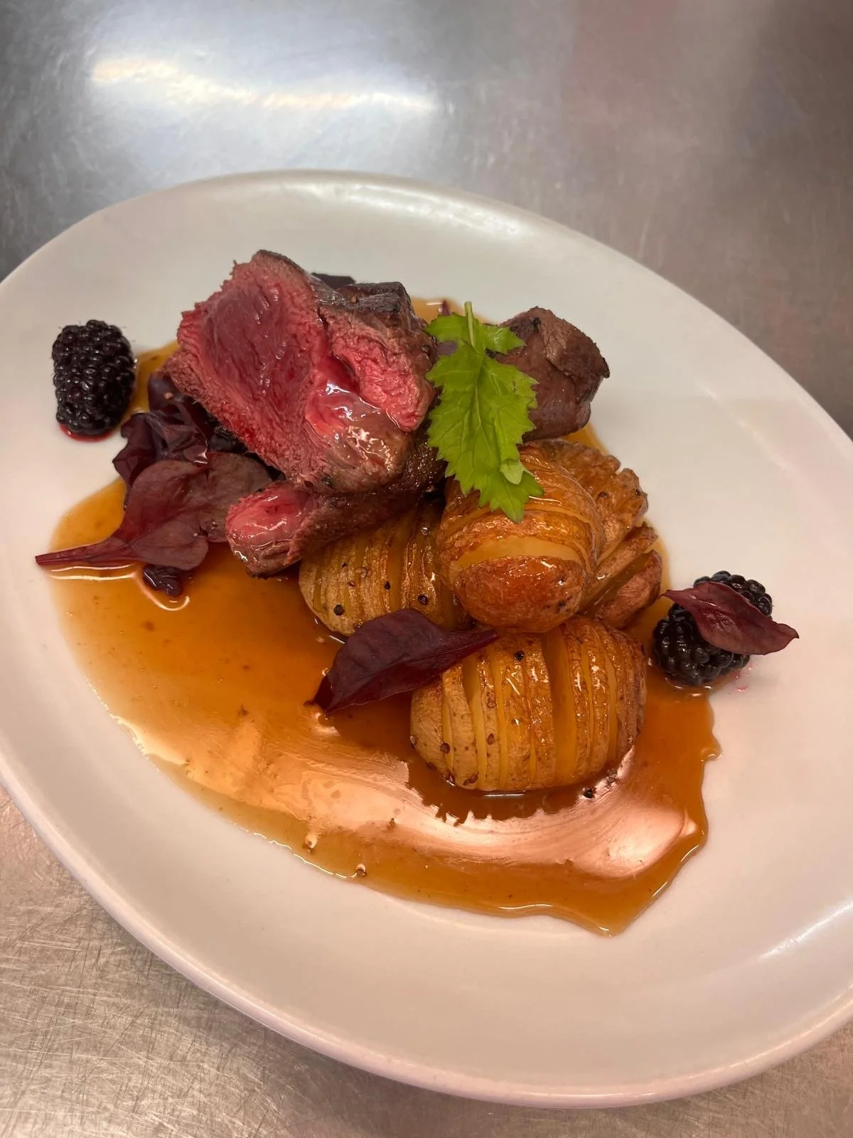 A plated dish with medium-rare steak, wrapped potatoes, and garnished with blackberries, purple leaves, and a green herb, in a light brown sauce.
