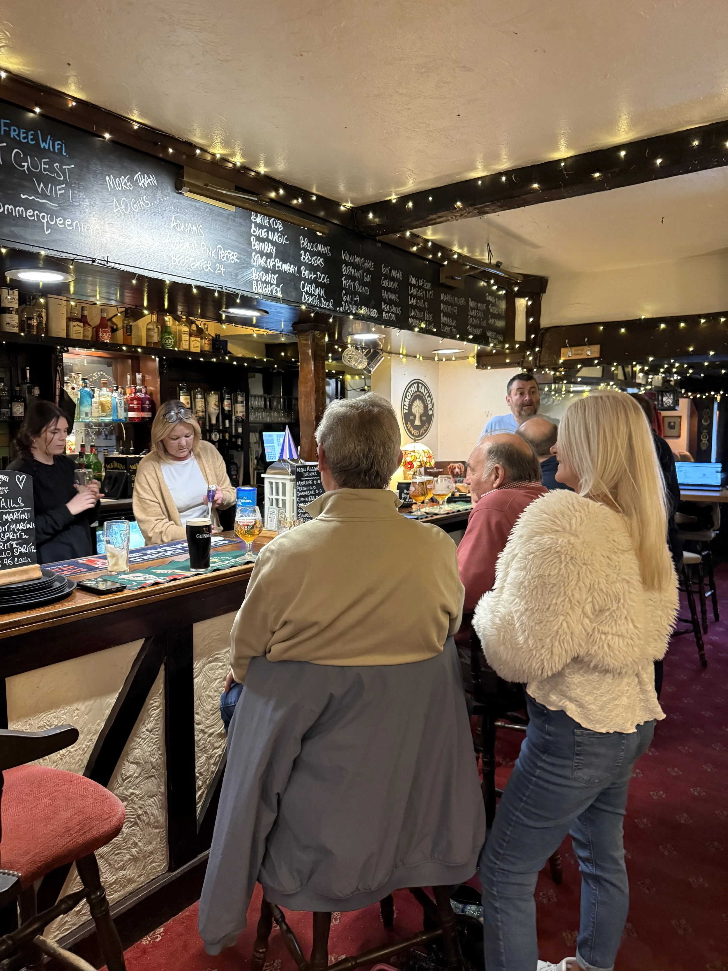 People sitting at a bar in a pub, ordering drinks, with a bartender serving, decorated with string lights and a chalkboard menu overhead.