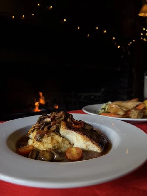 A plate of fish topped with mushrooms and sauce, served with mashed potatoes and vegetables in the background, in a dimly lit restaurant setting.
