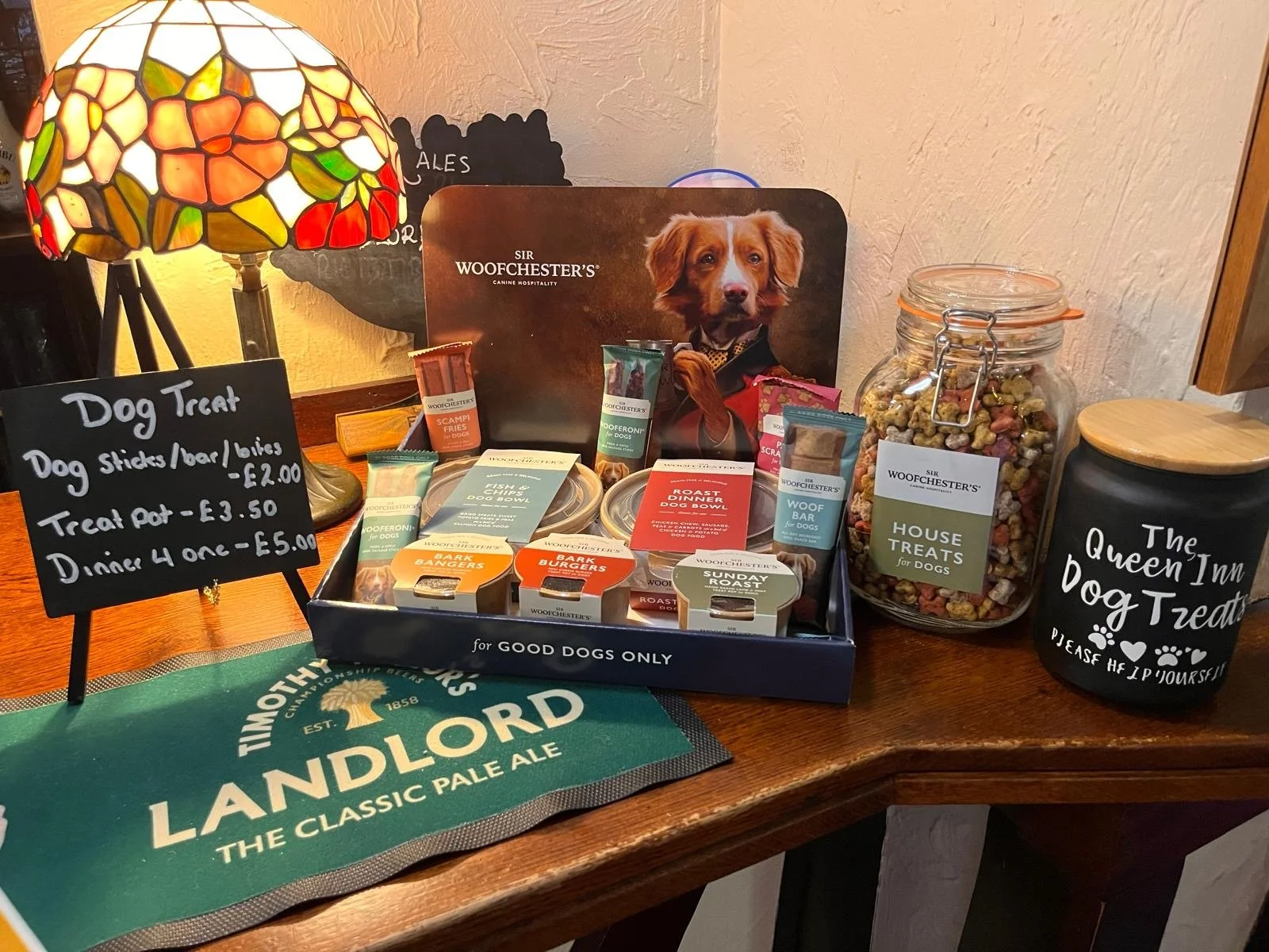 Display of dog treats and food items on a wooden table, including jars and boxes, with signs indicating prices and labels, a decorative lamp, and a mat with the Landlord branding.