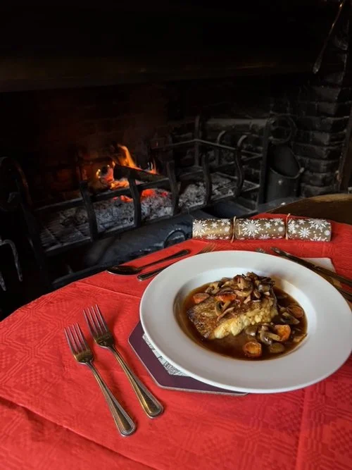 A plate of cooked fish with sauce and vegetables on a red tablecloth, set in front of a fireplace with burning logs.