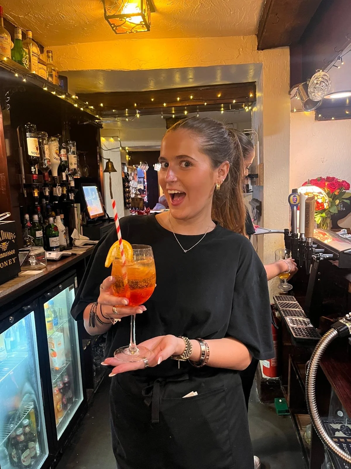 A young woman behind a bar holding a cocktail with an orange slice and striped straw, smiling at the camera. The bar is decorated with string lights and bottles of alcohol.