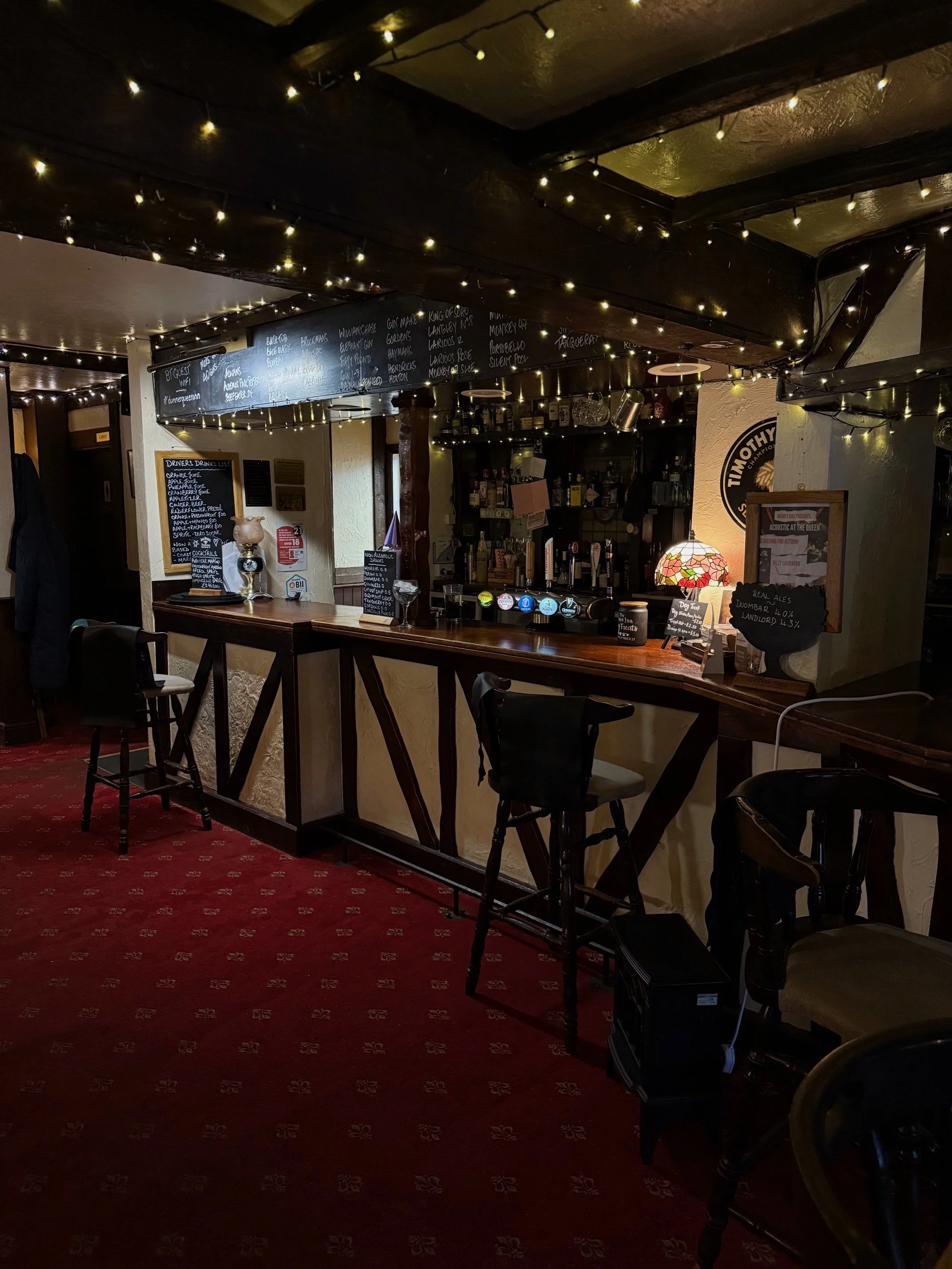 Inside a cozy pub with a wooden bar counter, adorned with string lights on the ceiling, a chalkboard menu, and bar stools. The background shows shelves with bottles and a small lamp on the bar.