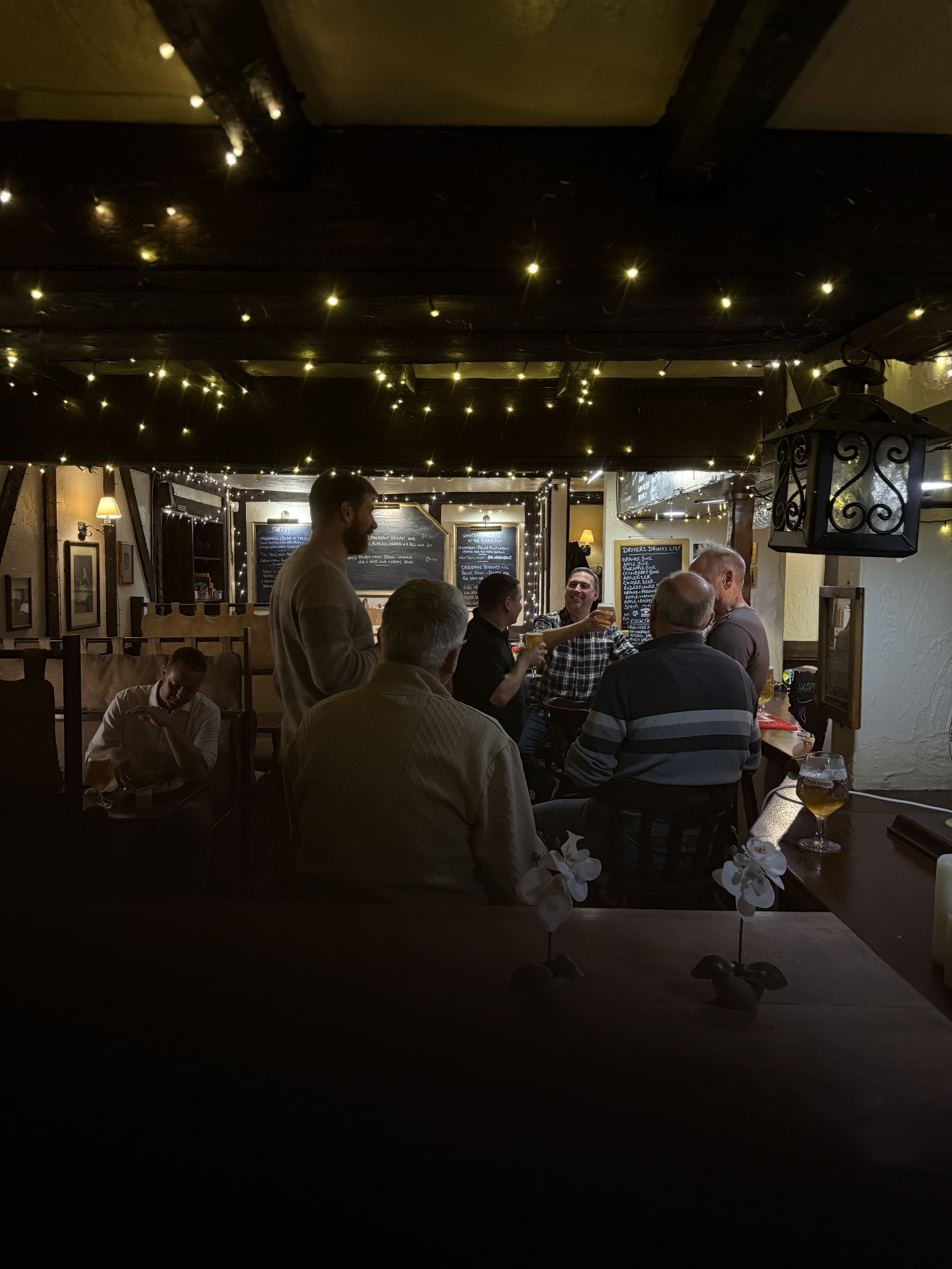Group of people sitting and standing in a cozy, dimly lit pub with string lights on the ceiling, several drinks on the tables, and chalkboards with menu items in the background.