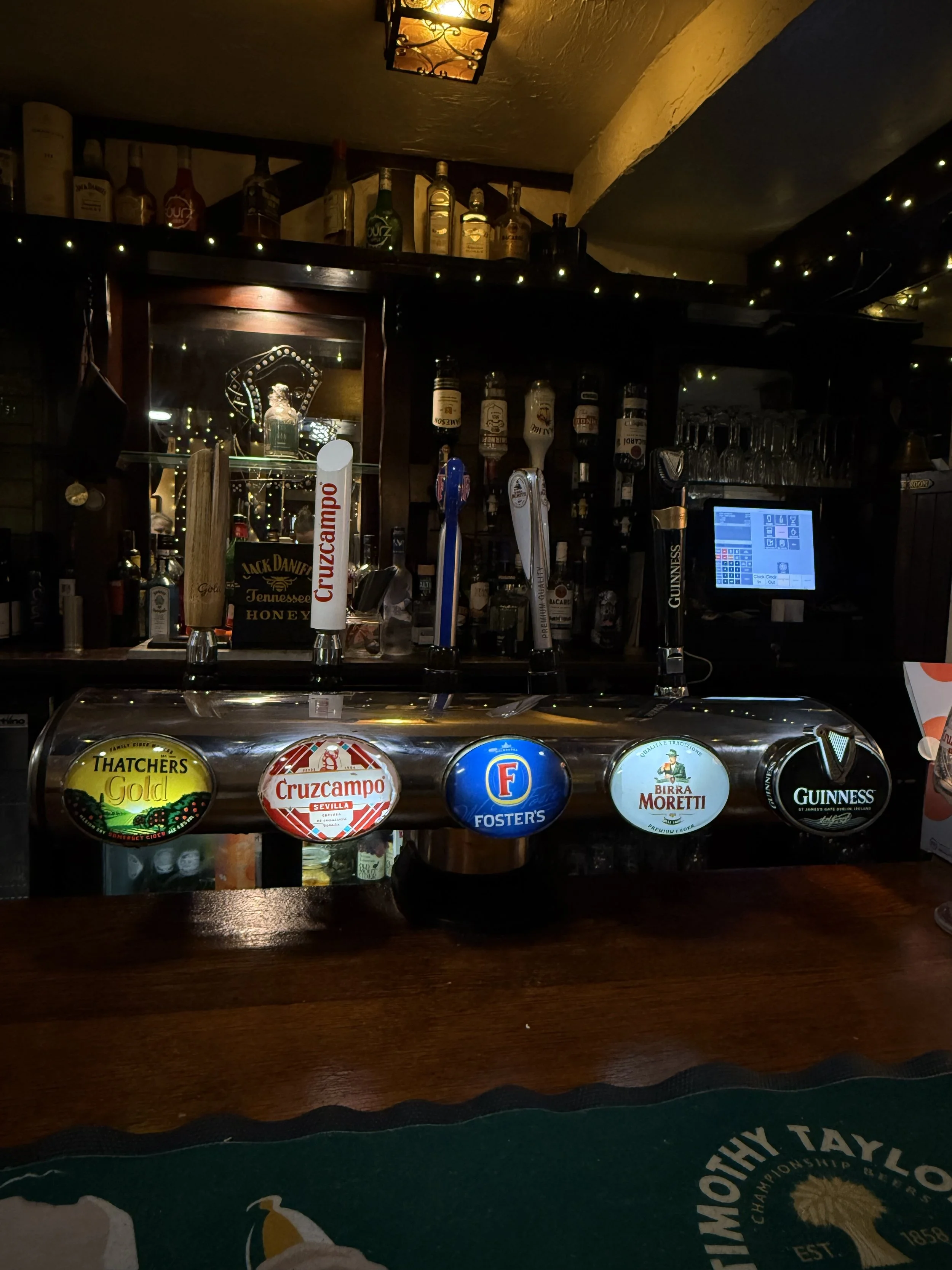 Bar with five beer tap handles displaying different beer brands, including Thatcher's Gold, Cruzcampo, Foster's, Birra Moretti, and Guinness, with a back bar stocked with liquor bottles and glasses, and a ceiling light fixture.