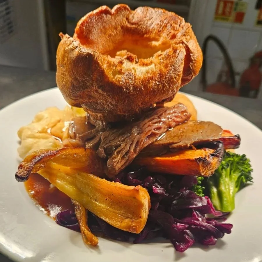 A plate of food with a bread bowl, roasted vegetables, and cooked meat with sides of mashed potatoes and broccoli.