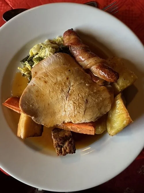 A plate of cooked food including a large piece of chicken, roasted potatoes, carrots, a bacon-wrapped item, and some cooked greens.