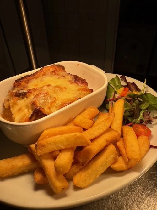 Baked dish with melted cheese served in a white bowl, side of french fries, and a small salad with greens and cherry tomatoes on a white plate.