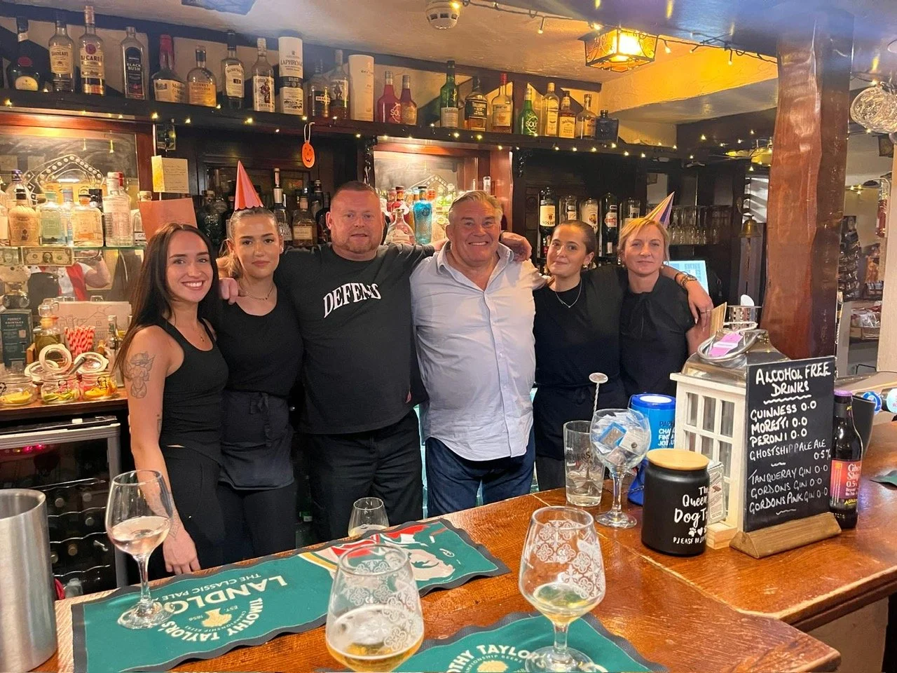 Group of six people celebrating at a bar, some wearing party hats, with drinks on the counter and bottles of alcohol behind them.