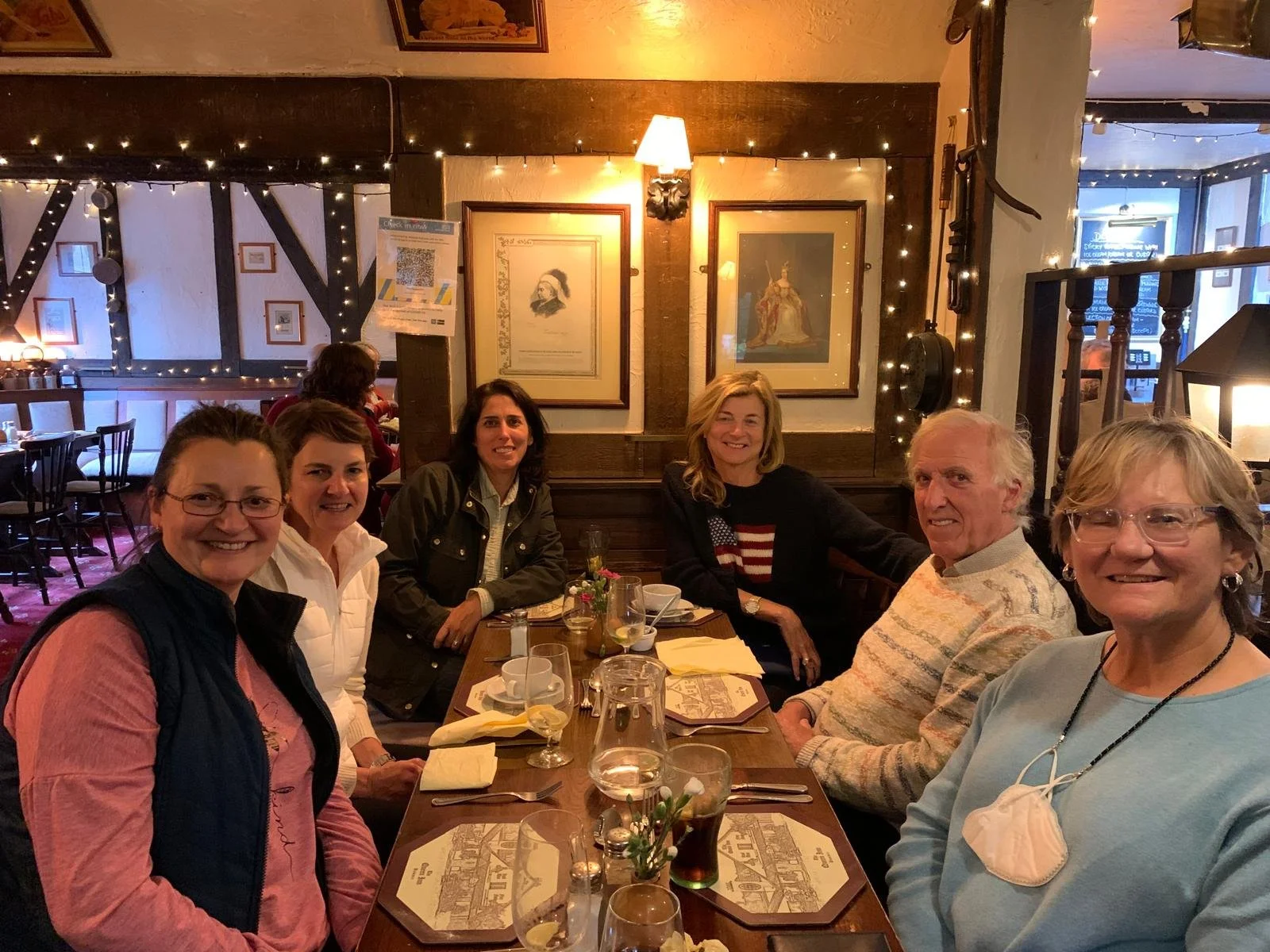 Seven adults sitting at a restaurant table, smiling for the camera. The table has glasses, napkins, and a small flower arrangement. The restaurant interior has framed artwork on the walls and fairy lights decorating the ceiling.