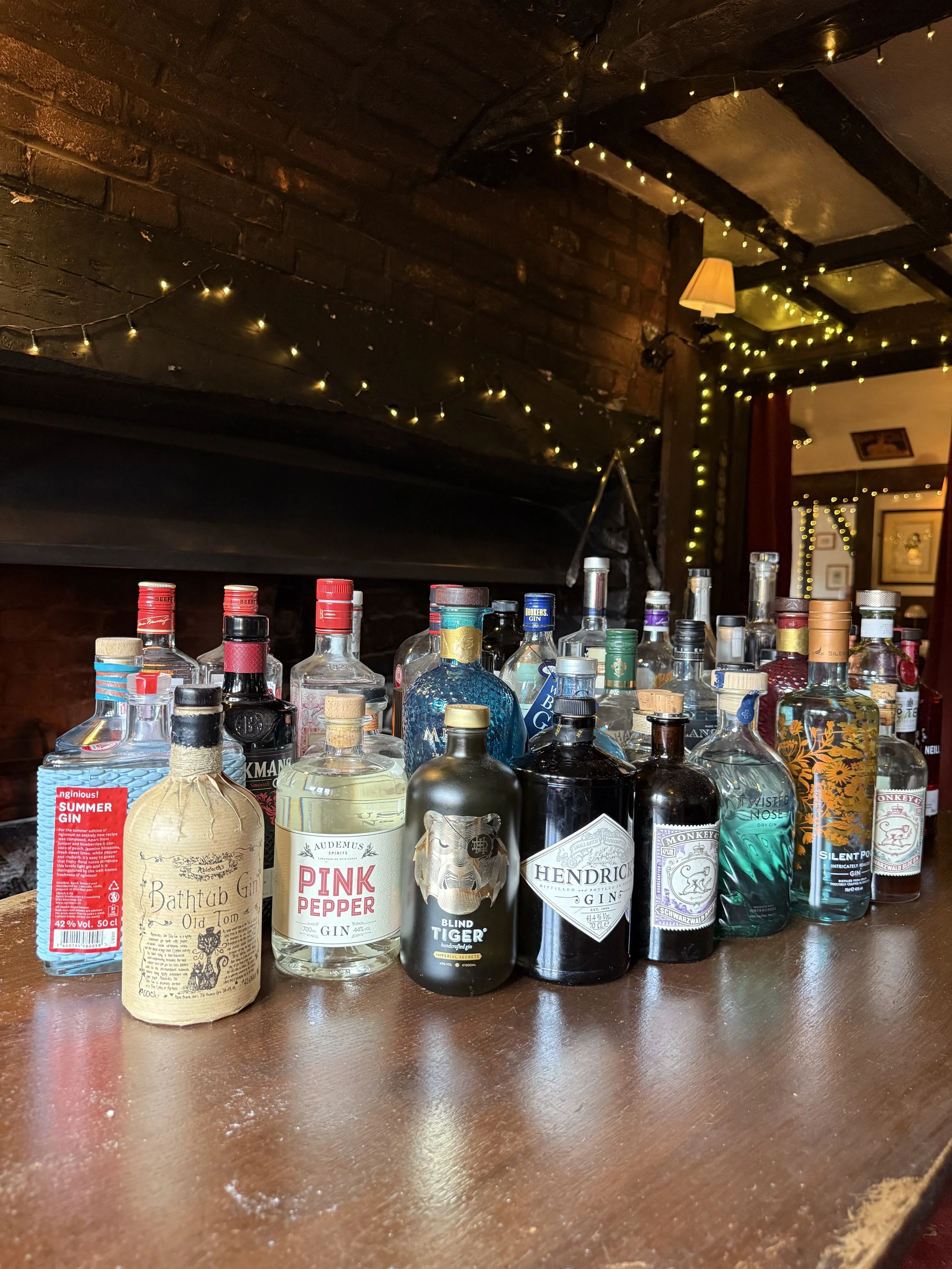 Collection of liquor bottles on a wooden bar with string and fairy lights, a dark brick wall, and warm ambient lighting.
