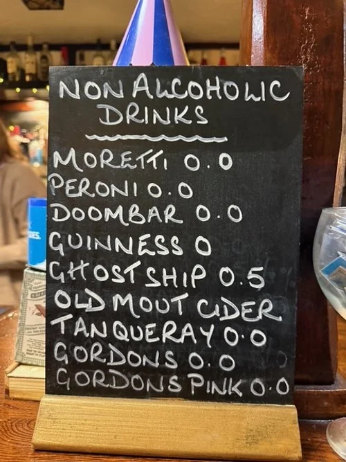 A chalkboard sign listing non-alcoholic drinks with prices, including Moretti, Peroni, Doombar, Guinness, Ghostship, Old Mout Cider, Tanqueray, Gordon's, and Gordon's Pink, with some priced at 0.0 and Ghostship at 0.5.