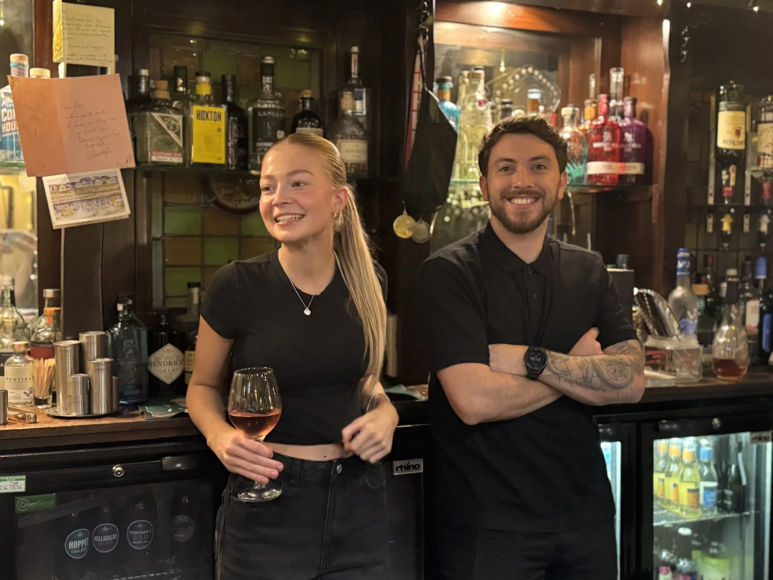 A woman with long blonde hair holding a glass of rosé wine, smiling and wearing a black crop top and black jeans, standing next to a smiling man with short dark hair and a beard, wearing a black shirt with tattoos on his arms, in a bar with shelves o