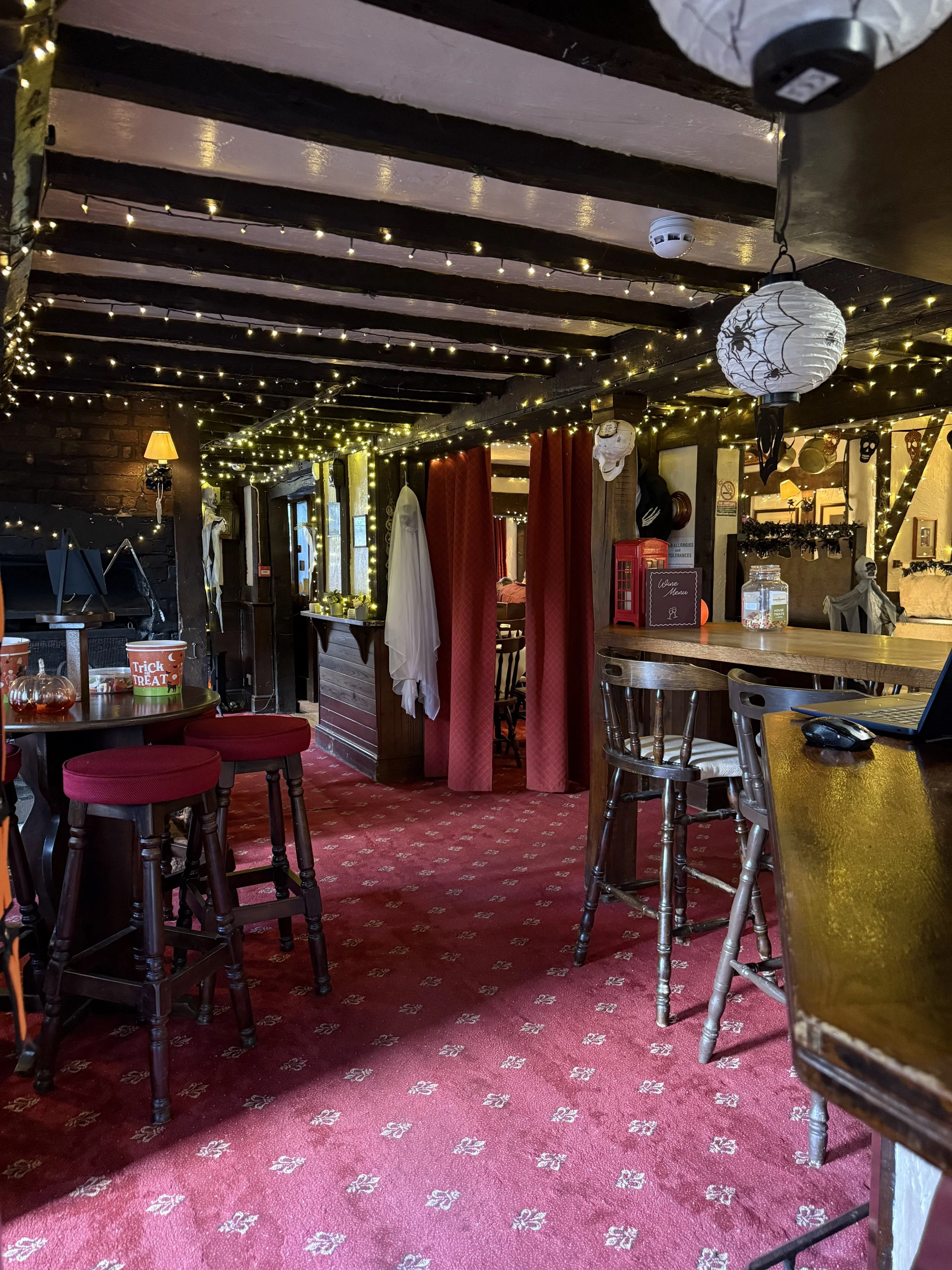 Interior of a cozy restaurant decorated with strings of fairy lights, halloween-themed paper lanterns, skeletons, and spider web decor, with red carpet, wooden furniture, and red curtains.