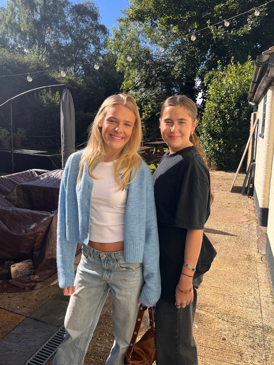 Two young women standing outdoors in a backyard with trees, string lights, and a building in the background. One is wearing a white crop top and light blue cardigan, and the other is in a black t-shirt. Both are smiling in sunlight.