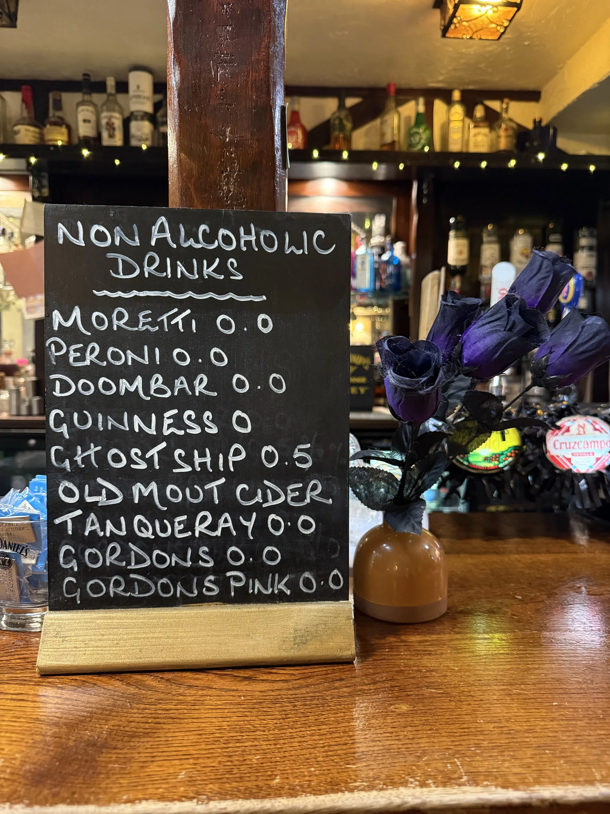A black chalkboard menu listing non-alcoholic drinks with names and prices, placed on a wooden bar counter next to a small vase with purple roses, inside a bar with bottles of alcohol in the background.
