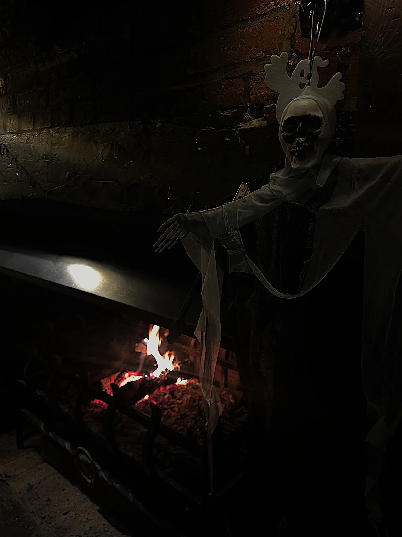 A Halloween decoration of a skeleton with a skull face wearing a hoodie, posed next to a fireplace with burning logs, against a dark brick wall.