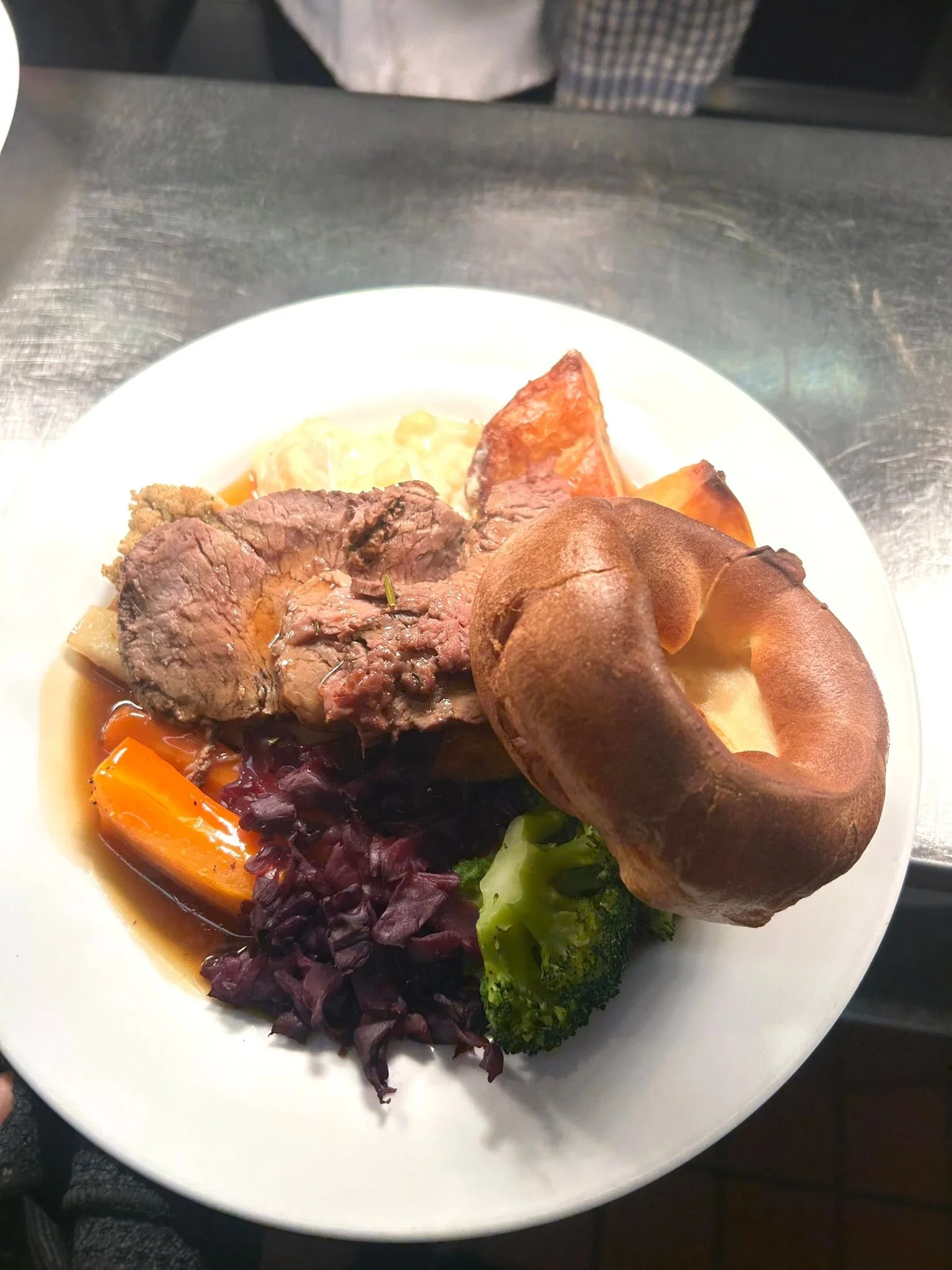 A plate of cooked roast beef served with roasted carrots, purple cabbage, broccoli, and a baked potato with a brown skin.
