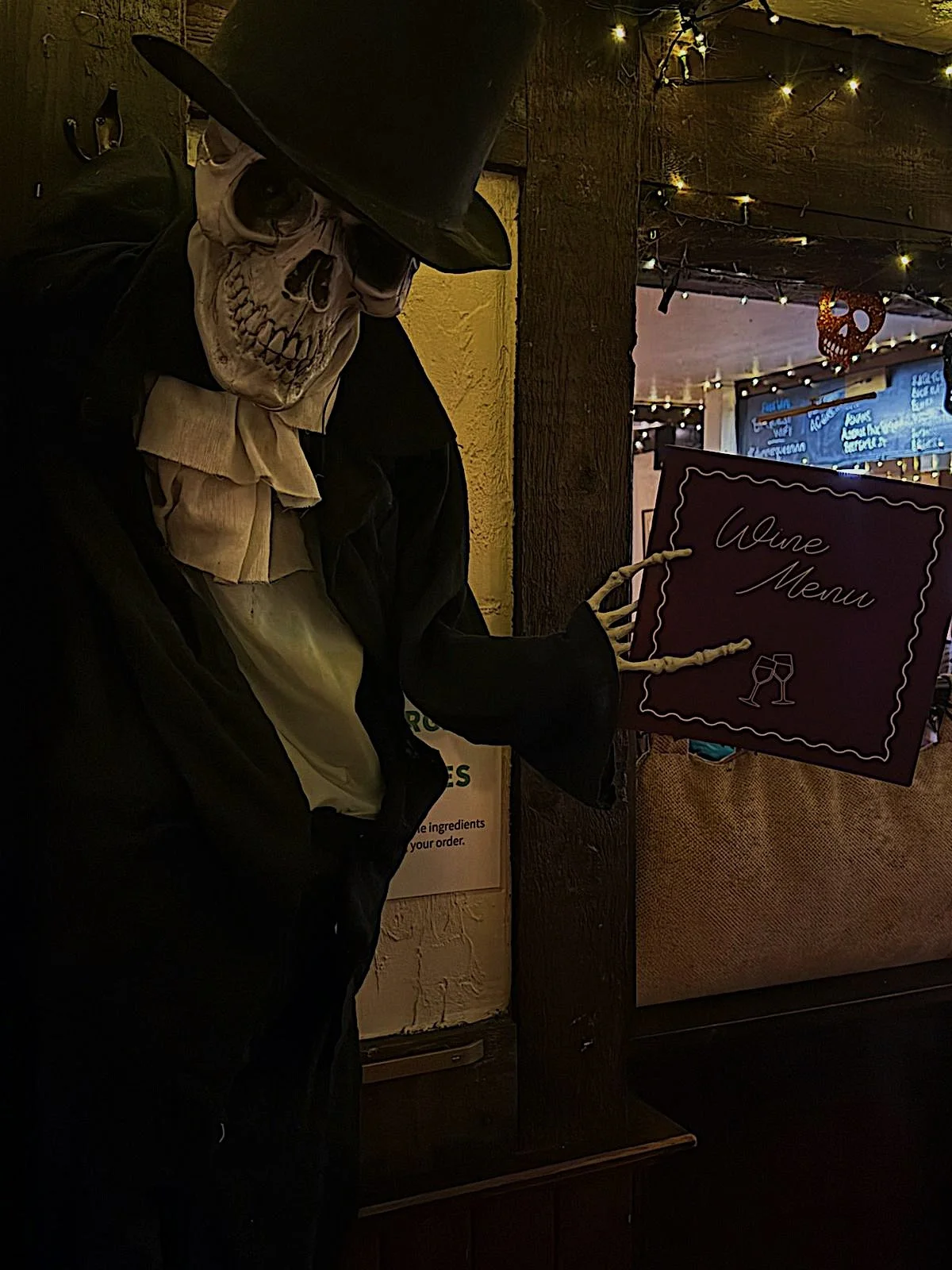 A skeleton dressed in a black coat and hat holding a wine menu at a dimly lit restaurant or bar decorated with fairy lights and Halloween ornaments.