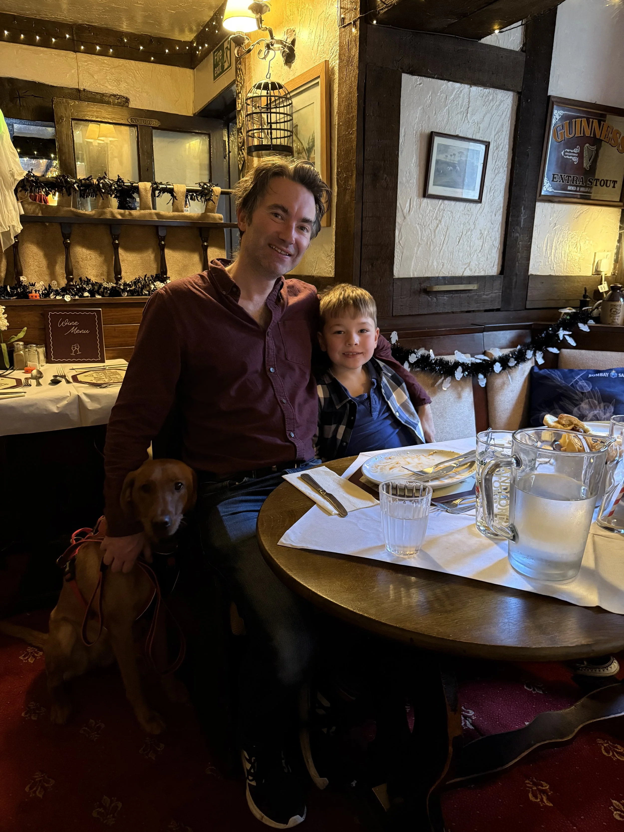 A man, a young boy, and a dog sitting at a dinner table in a cozy restaurant with warm lighting and festive decorations.